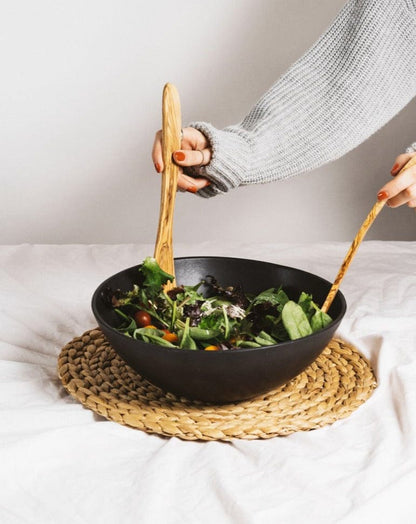 Person holding a black bowl with salad on a woven mat, The Salad Essential brings together Salad Bowl and Forks.