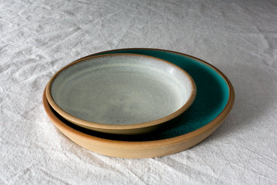 Pale grey-green Settle Ceramics Fat Rim Lunch Plate resting on a larger deep teal plate over textured white cloth, showing contrasting brown edges and softly speckled ceramic glaze.