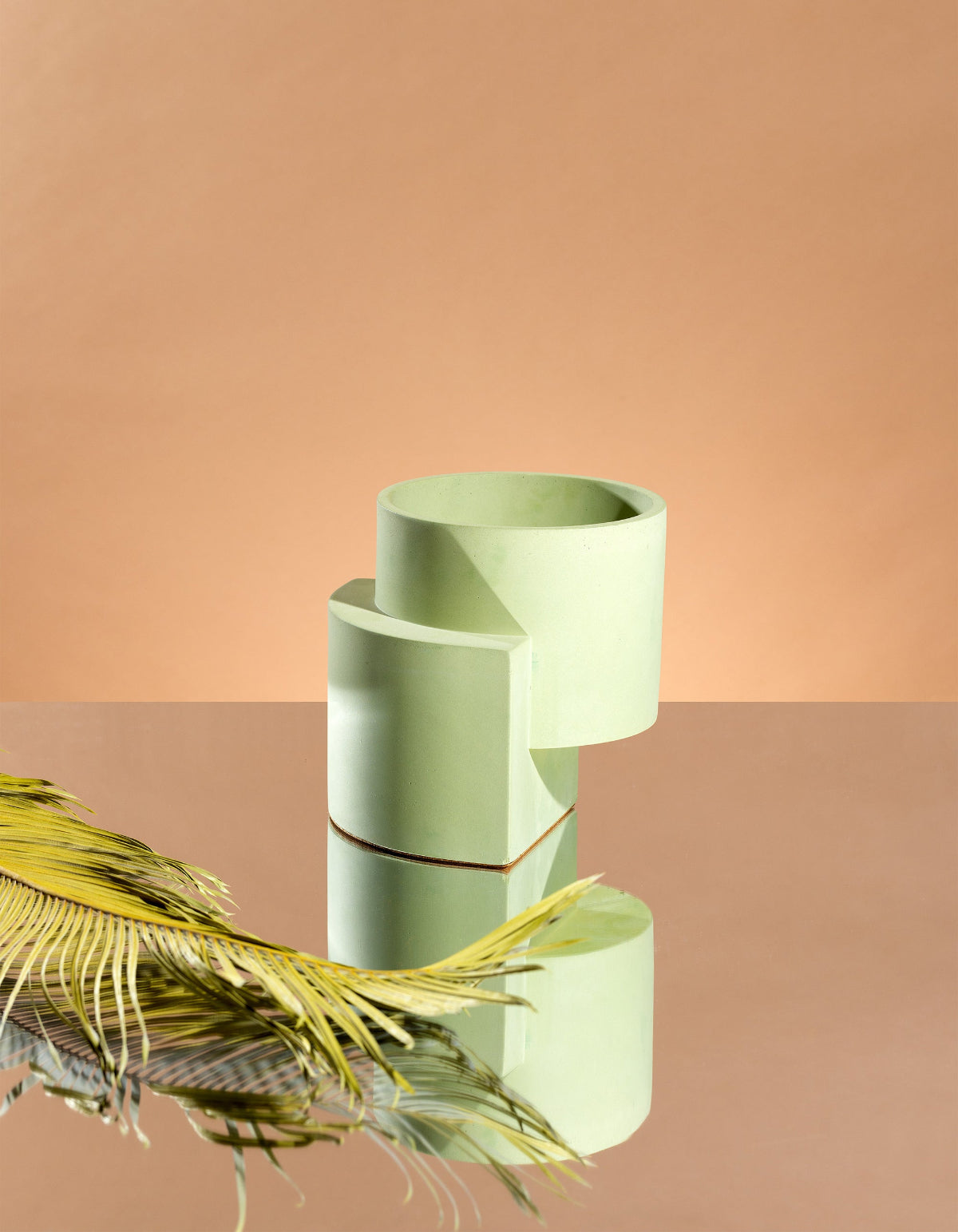 Pale green Tortuga Forma Platform Planter Medium - Polished Concrete on a mirrored surface against a peach backdrop, with dried stems beside it and a reflection highlighting its stepped geometric shape.