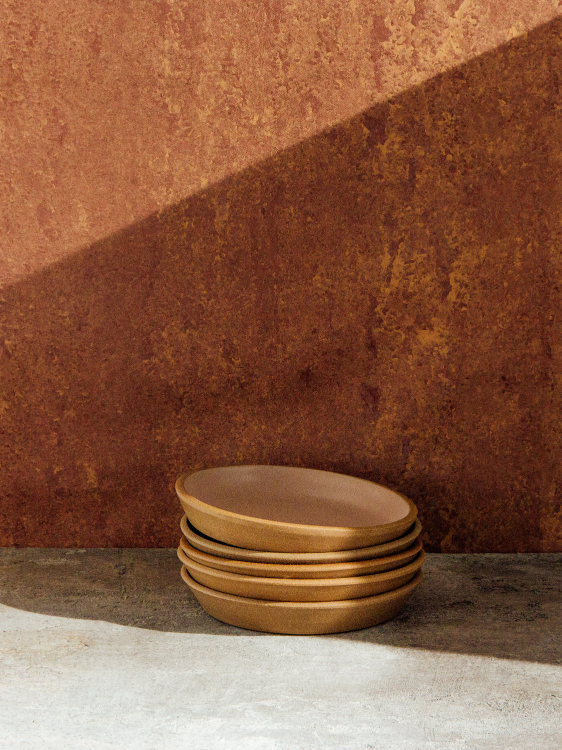 Stack of golden-brown Settle Ceramics Fat Rim Lunch Plates on a pale ledge against a rust-coloured wall, with thick rounded edges highlighted beneath a diagonal band of sunlight.