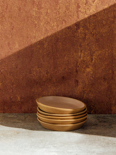 Stack of golden-brown Settle Ceramics Fat Rim Lunch Plates on a pale ledge against a rust-coloured wall, with thick rounded edges highlighted beneath a diagonal band of sunlight.