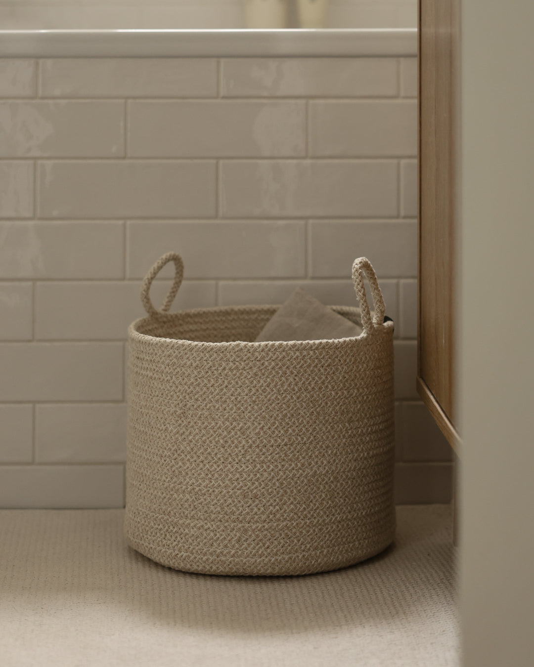 Large beige rope basket holding folded textiles beside a tiled wall.