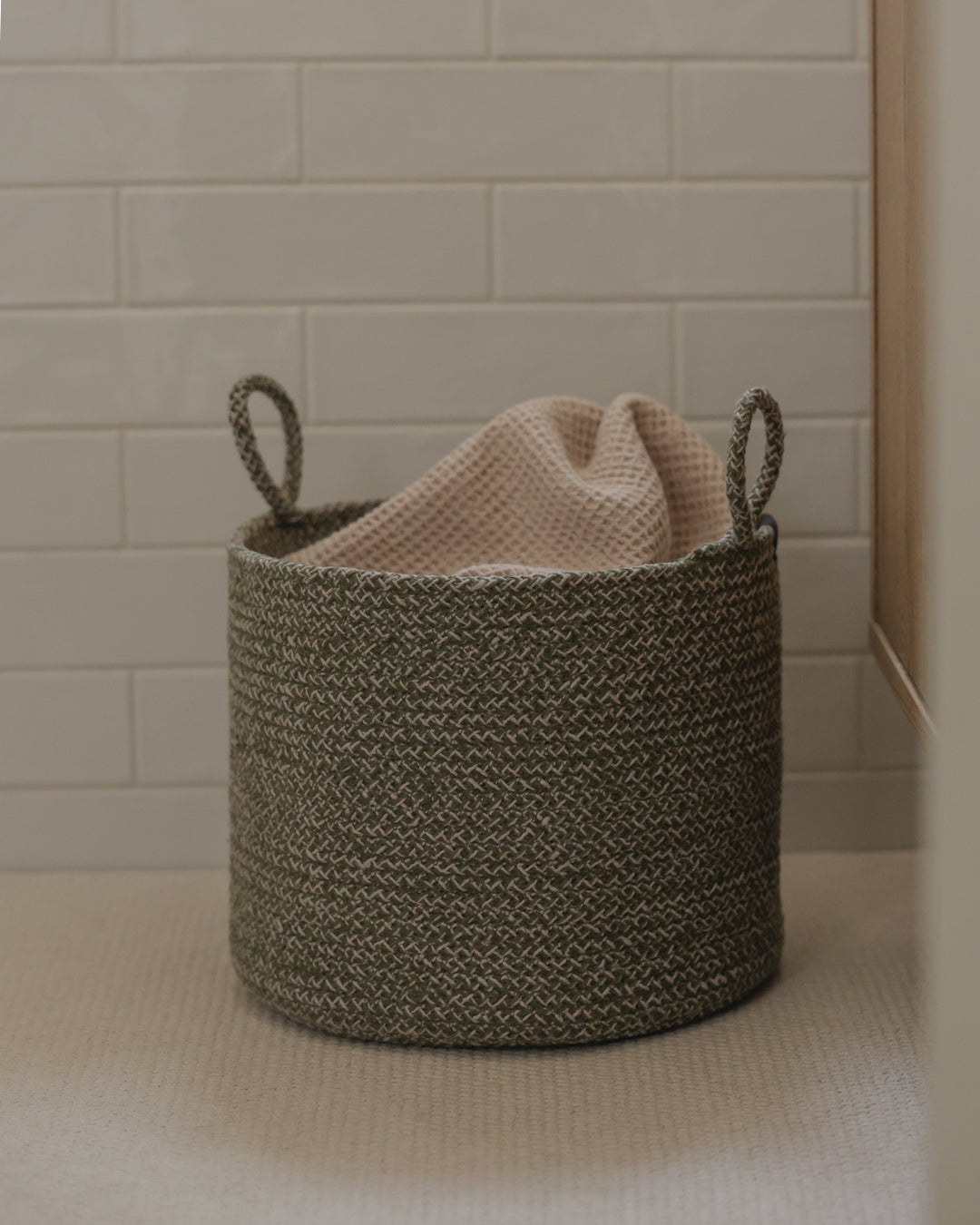 Green melange basket holding a soft beige blanket beside a tiled wall.