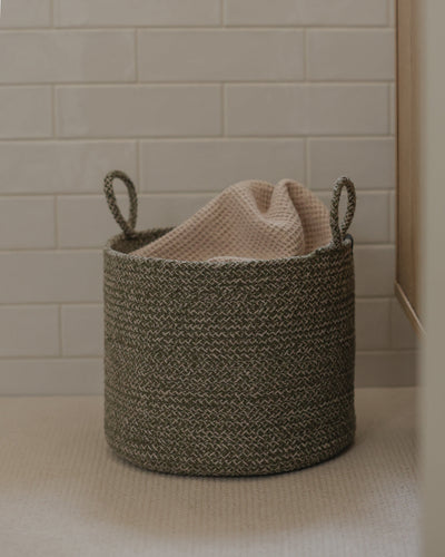Green melange basket holding a soft beige blanket beside a tiled wall.
