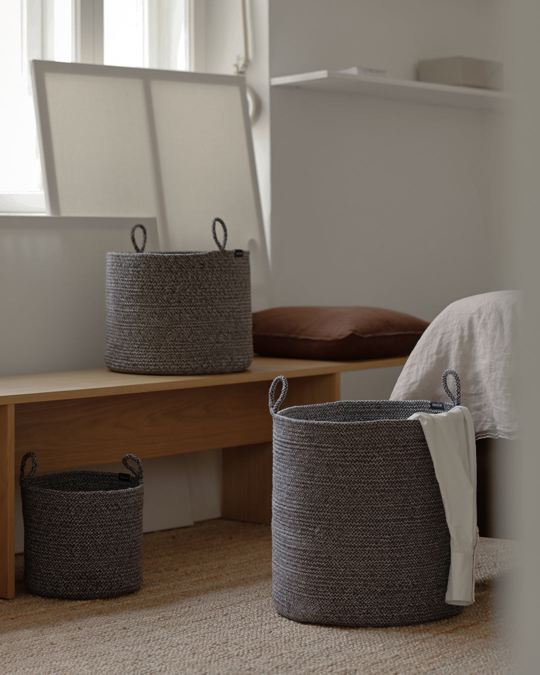 Three basket sizes arranged around a wooden bench in a softly lit bedroom.