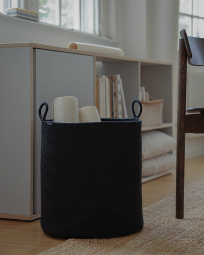 Large black rope basket holding rolled textiles beside a shelving unit in a modern room.