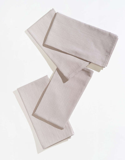 Four pale grey Tortuga Forma Dinner Napkins Sabi folded into overlapping rectangles on white, showing fine linear woven texture, stitched hems and soft shadows beneath the edges.