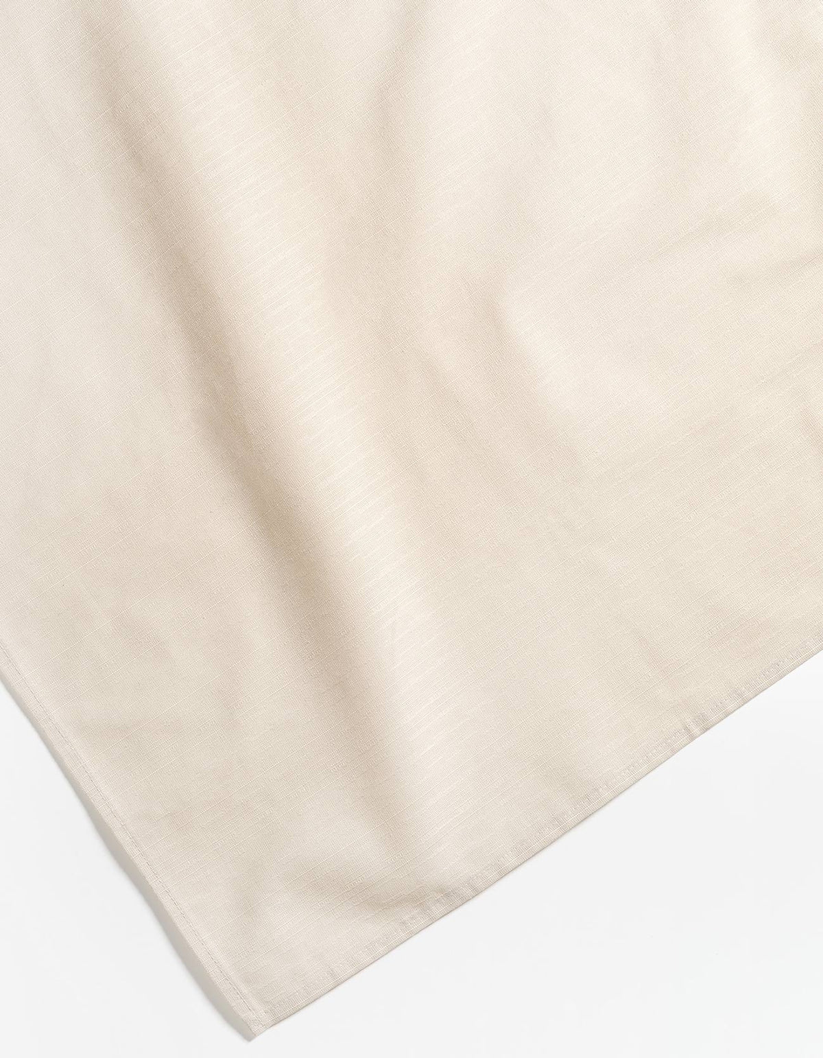 Close-up of an ivory Tortuga Forma Tablecloth Sabi corner against white, showing its subtle woven texture, gentle creases and neatly stitched hem.