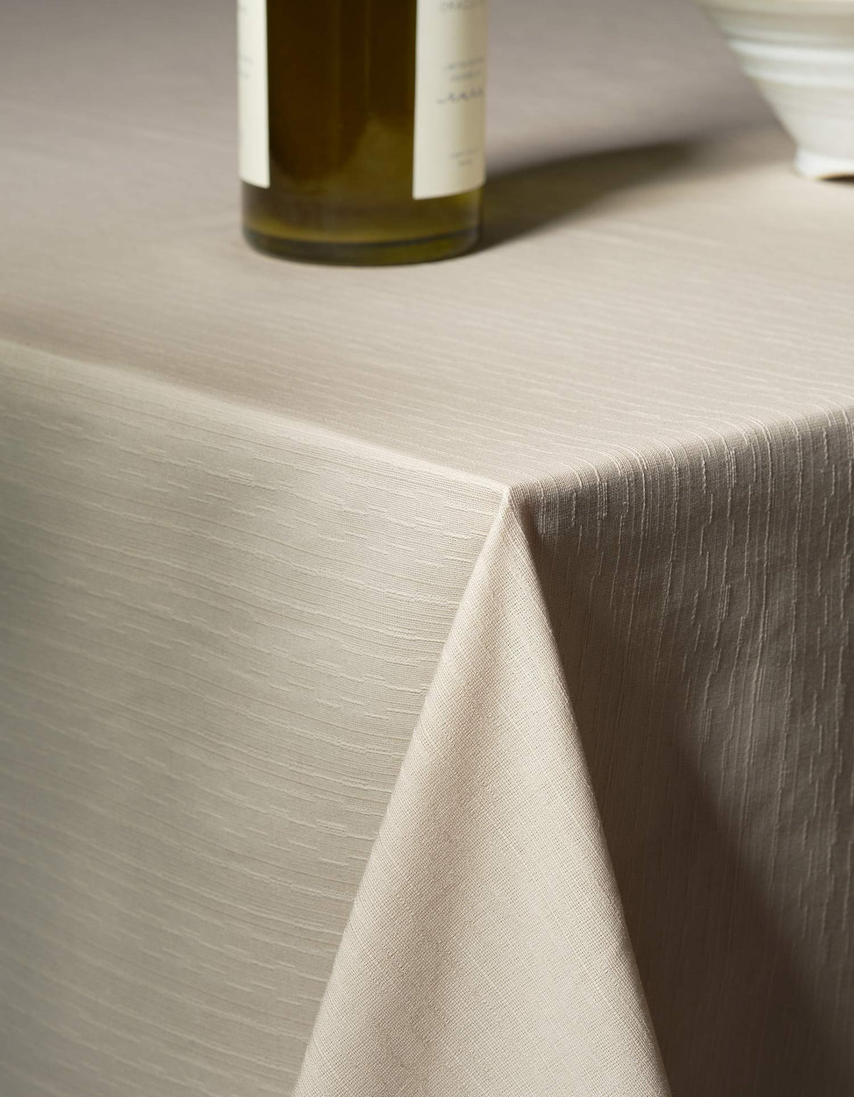 Ivory Tortuga Forma Tablecloth Sabi draped over a table corner, showing fine linear woven texture and soft vertical folds, with a green glass bottle and part of a white bowl on top.