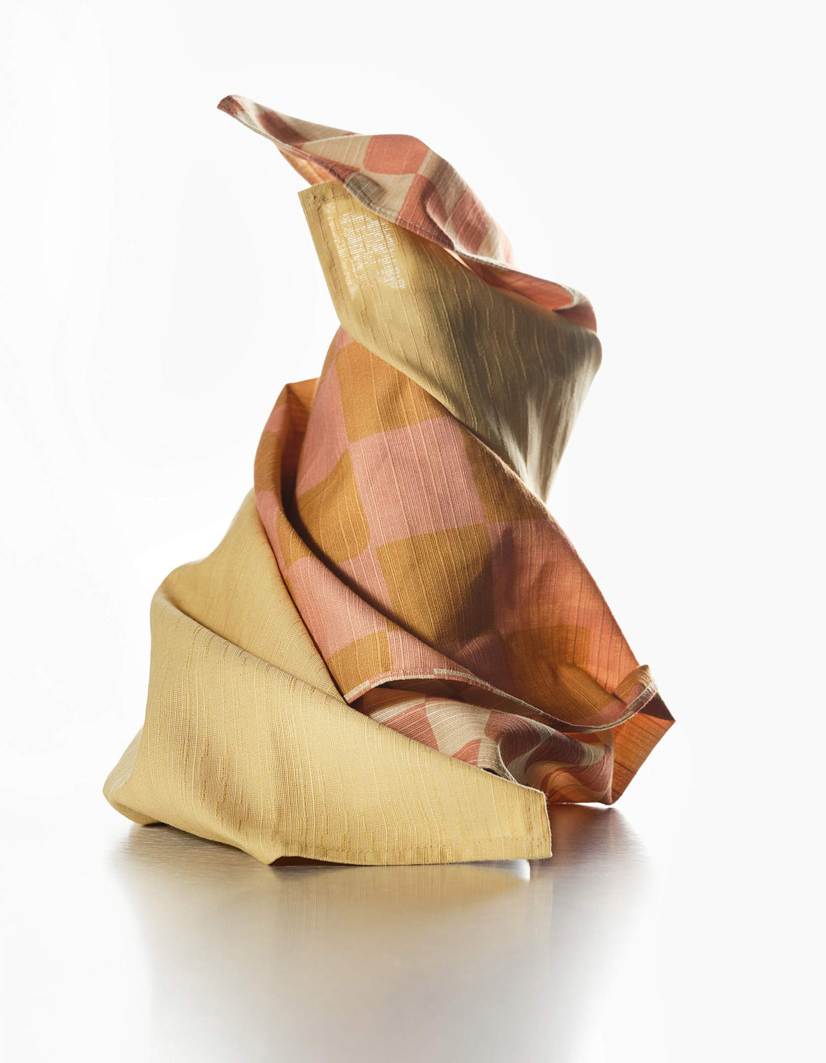 Mustard-yellow Tortuga Forma Dinner Napkins Sabi gathered into sculptural folds with rust, pink and olive patterned fabric, showing the plain napkin’s texture and stitched edge.