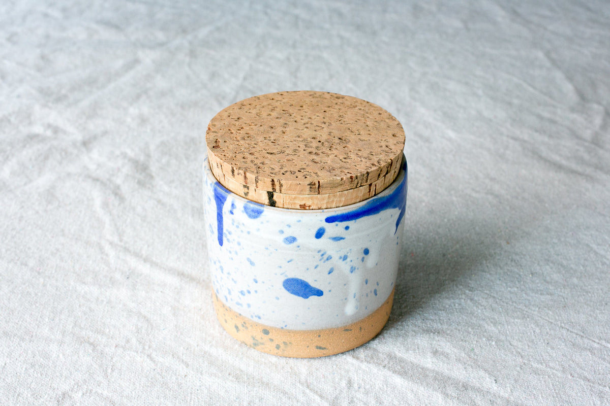 White Settle Ceramics Wander Salt Jar with blue splashes and a blue band around the rim, topped with a round cork lid and finished with a tan base, standing on pale textured fabric.