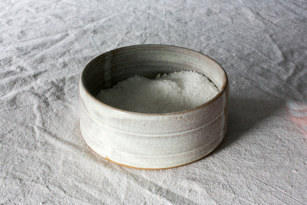 Pale grey-green Settle Ceramics Wander Salt Pig filled with white salt on wrinkled light grey fabric, viewed from above at an angle, showing its open cylindrical ceramic body, horizontal ridges, darker interior and narrow ochre base.