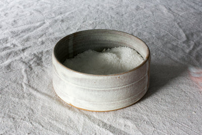 Pale grey-green Settle Ceramics Wander Salt Pig filled with white salt on wrinkled light grey fabric, viewed from above at an angle, showing its open cylindrical ceramic body, horizontal ridges, darker interior and narrow ochre base.