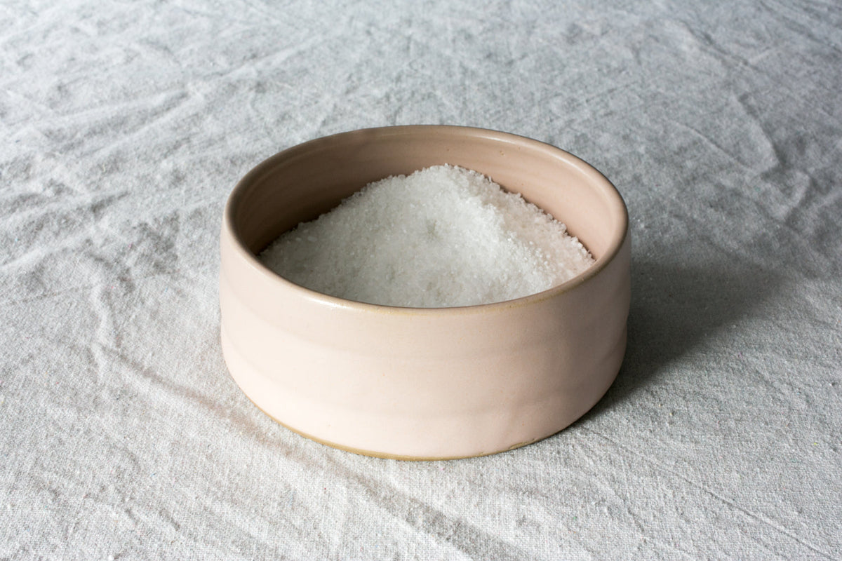 Cream Settle Ceramics Wander Salt Pig holding a mound of white salt on light grey fabric, showing its low round shape, open top, warm beige interior and subtle horizontal surface lines.
