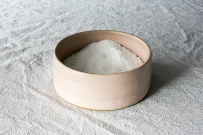 Cream Settle Ceramics Wander Salt Pig holding a mound of white salt on light grey fabric, showing its low round shape, open top, warm beige interior and subtle horizontal surface lines.