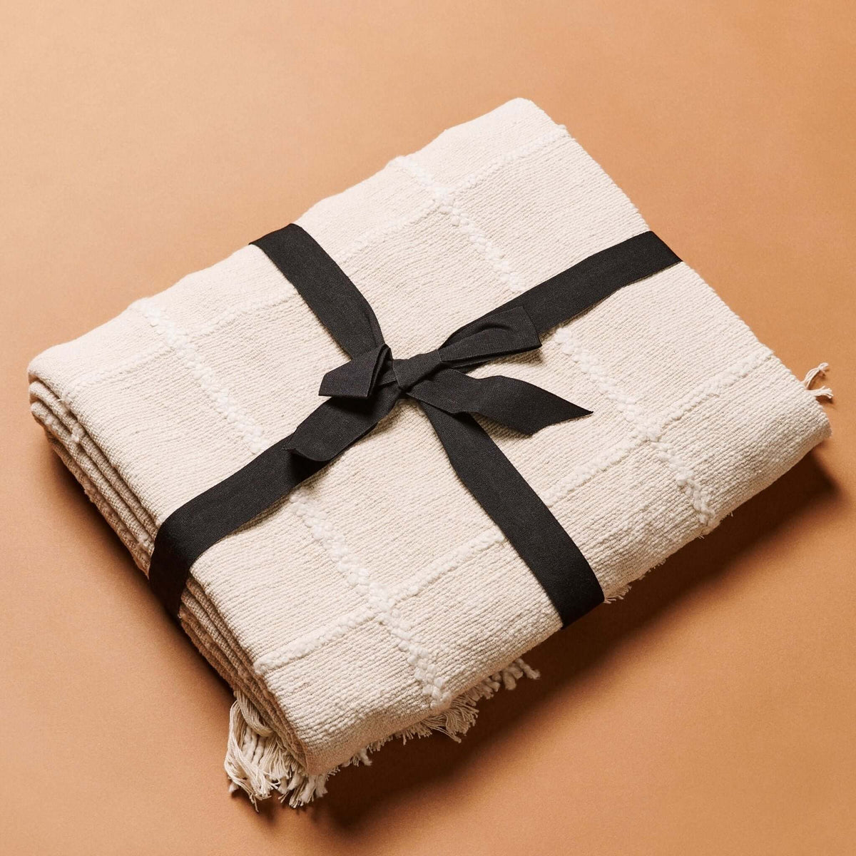 Folded Sea Shell White Textured Checks Handloom Throw wrapped with a black ribbon against a warm tan background