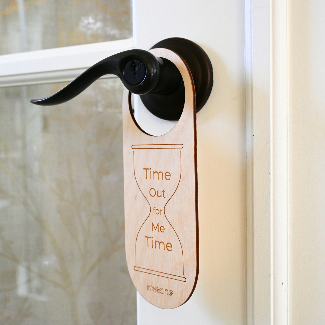 Light natural wood door hanger suspended from a black lever handle, engraved with an hourglass outline and the words “Time Out for Me Time.”