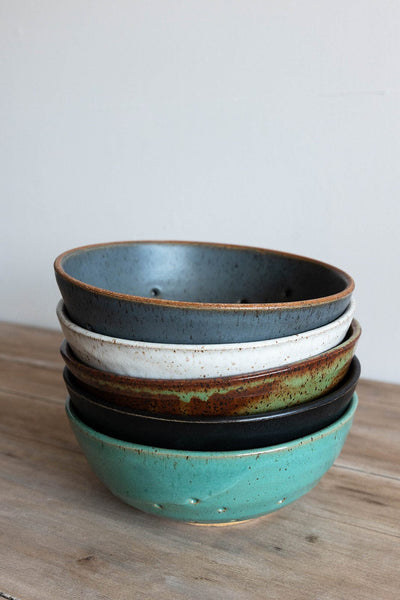 Stack of ceramic colander bowls with different colors on a wooden surface