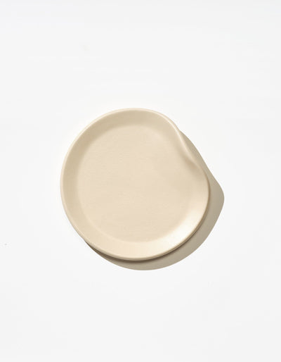 Tortuga Forma Bread Plate Single in Bone viewed overhead on white, showing a softly asymmetrical round shape, shallow raised rim and inward curve along one side, with subtle texture across its glazed surface.