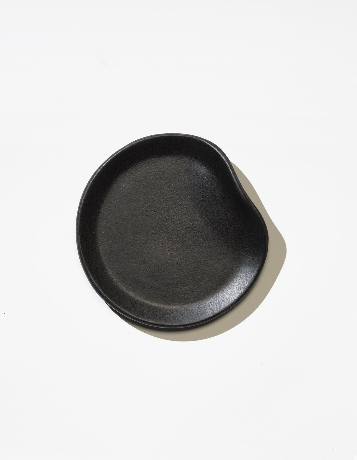 Tortuga Forma Bread Plate Single in Soot viewed overhead on white, showing a shallow centre, fine surface texture and raised rim with an inward curve on one side.