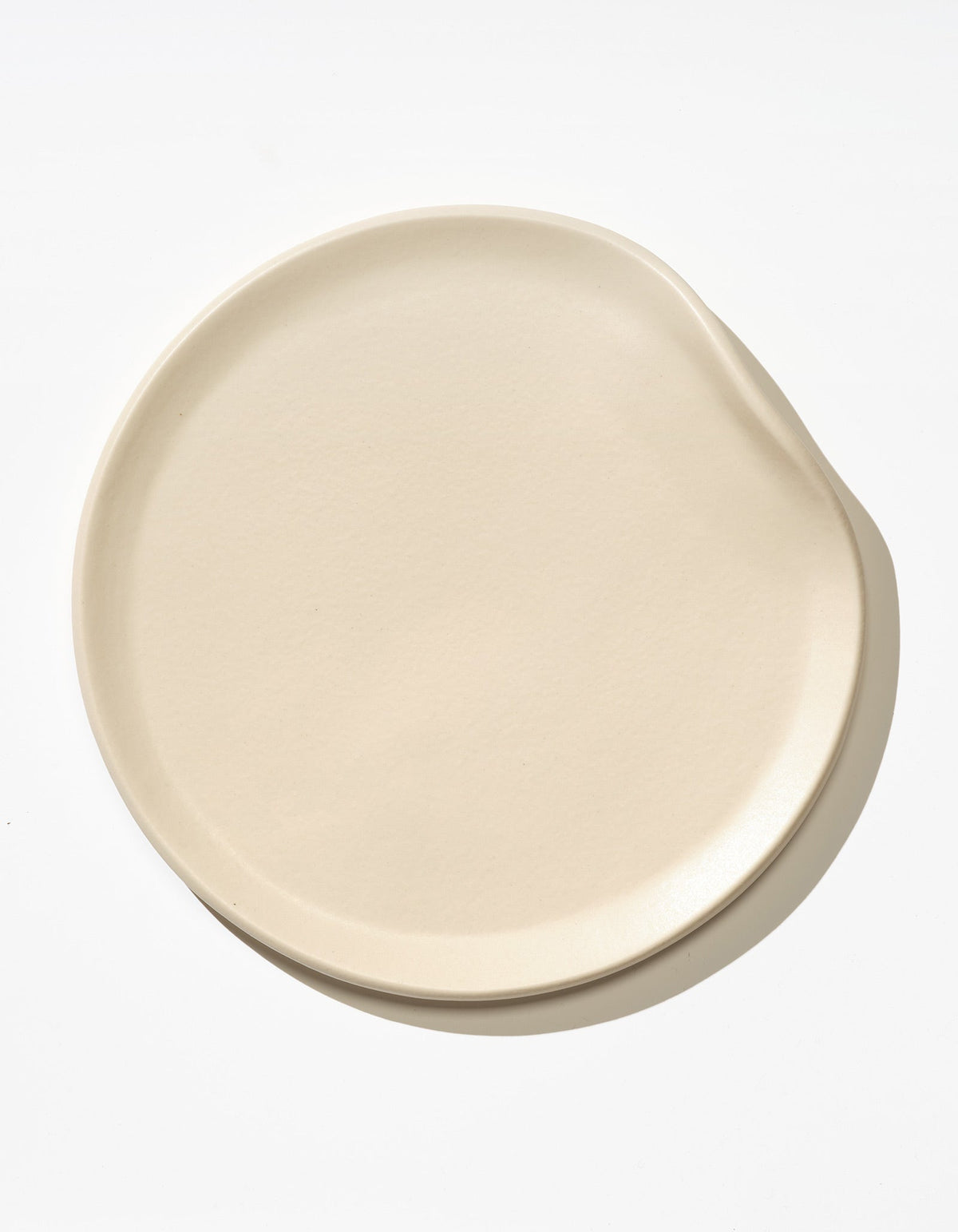 Tortuga Forma Dinner Plate Single in Bone viewed overhead on white, showing a softly asymmetrical round shape, shallow raised rim and smooth glazed surface with subtle texture.