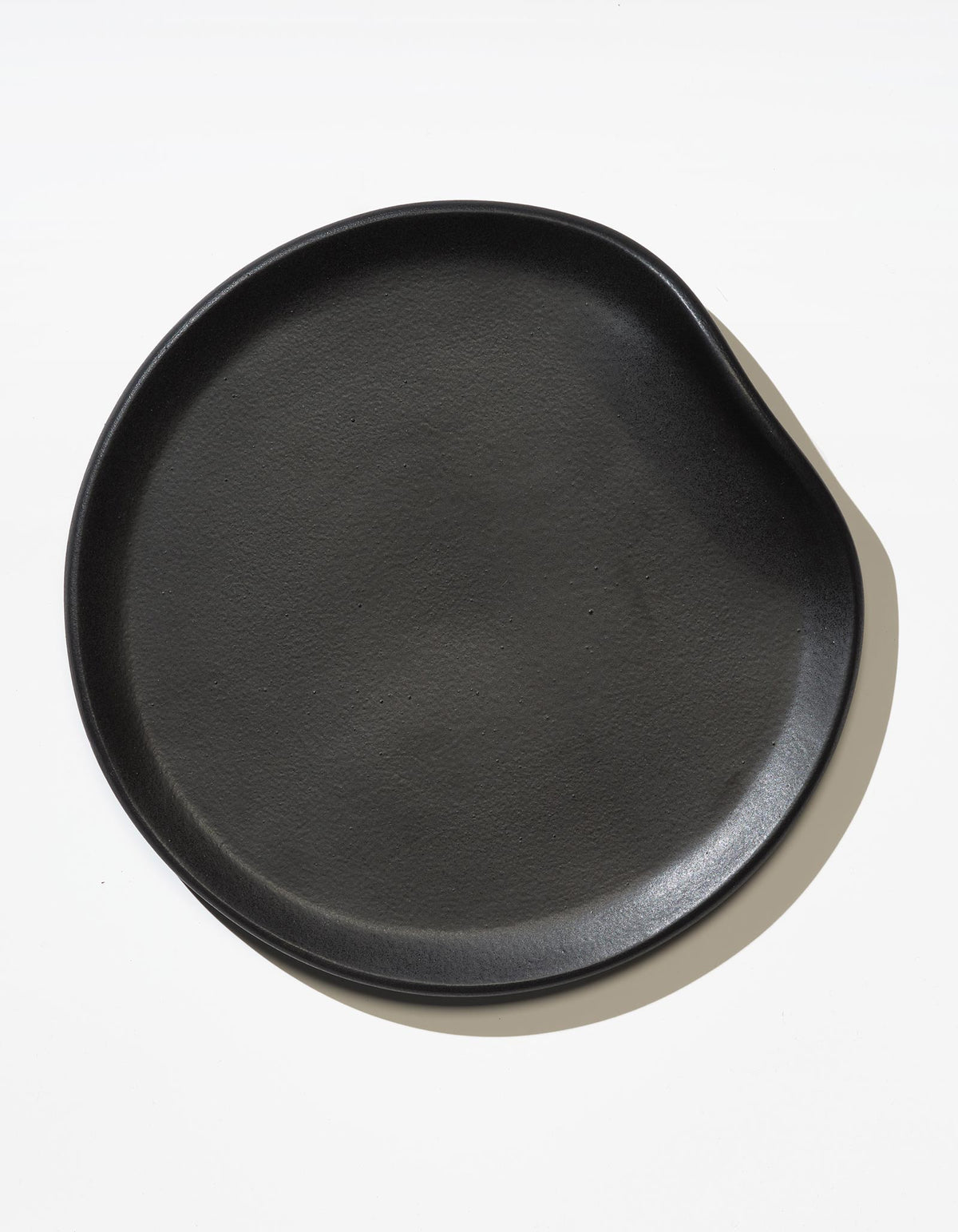 Tortuga Forma Dinner Plate Single in Soot viewed overhead on white, showing a softly asymmetrical circular form, shallow rim and fine surface texture highlighted by side lighting.