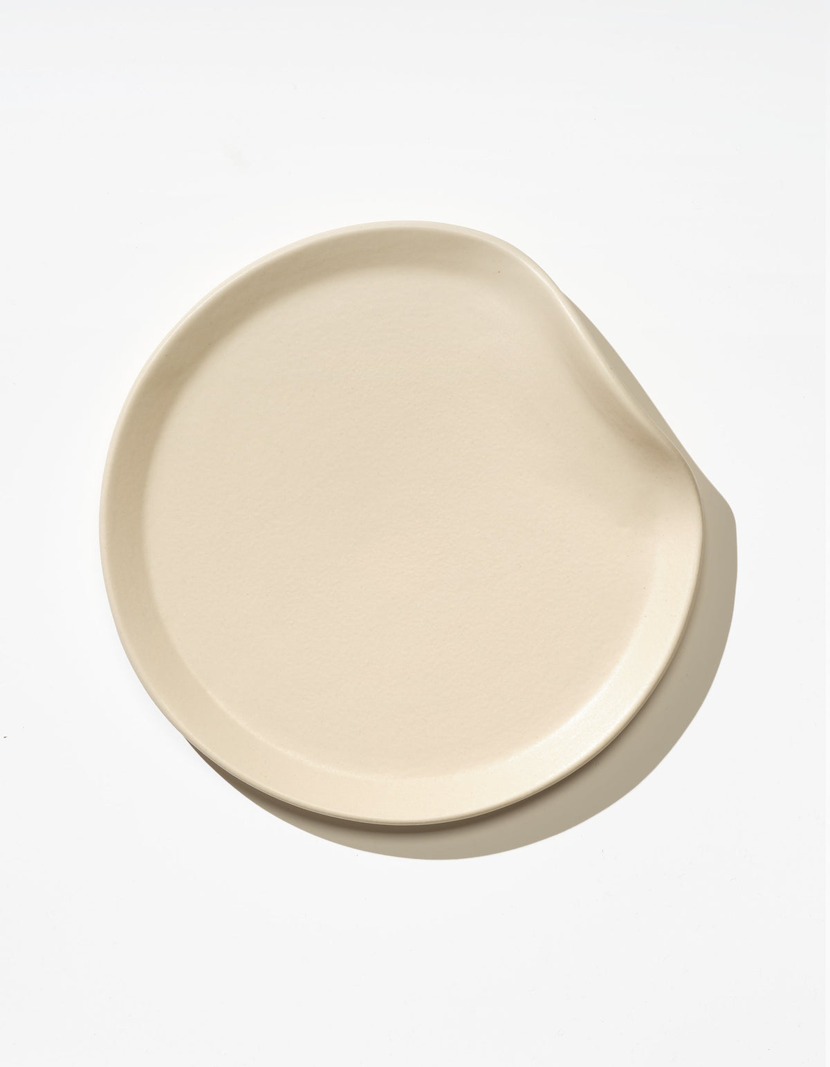 Tortuga Forma Salad Plate Single in Bone viewed overhead on white, showing a softly irregular round shape, shallow raised rim and gently indented edge, with light reflecting across its glazed surface.
