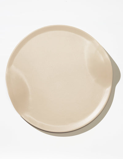 Tortuga Forma Serve Platter in Bone off-white viewed overhead on white, showing a broad round centre, shallow raised rim and two opposing inward curves, with light reflecting across its smooth glazed surface.