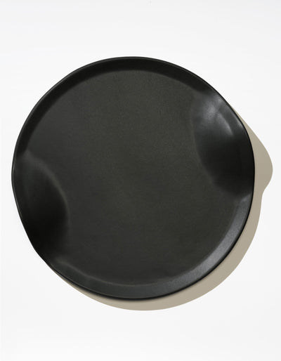 Tortuga Forma Serve Platter in Soot black viewed overhead on white, showing a broad serving surface, low raised rim and two opposing inward curves, with reflected light highlighting the smooth finish.