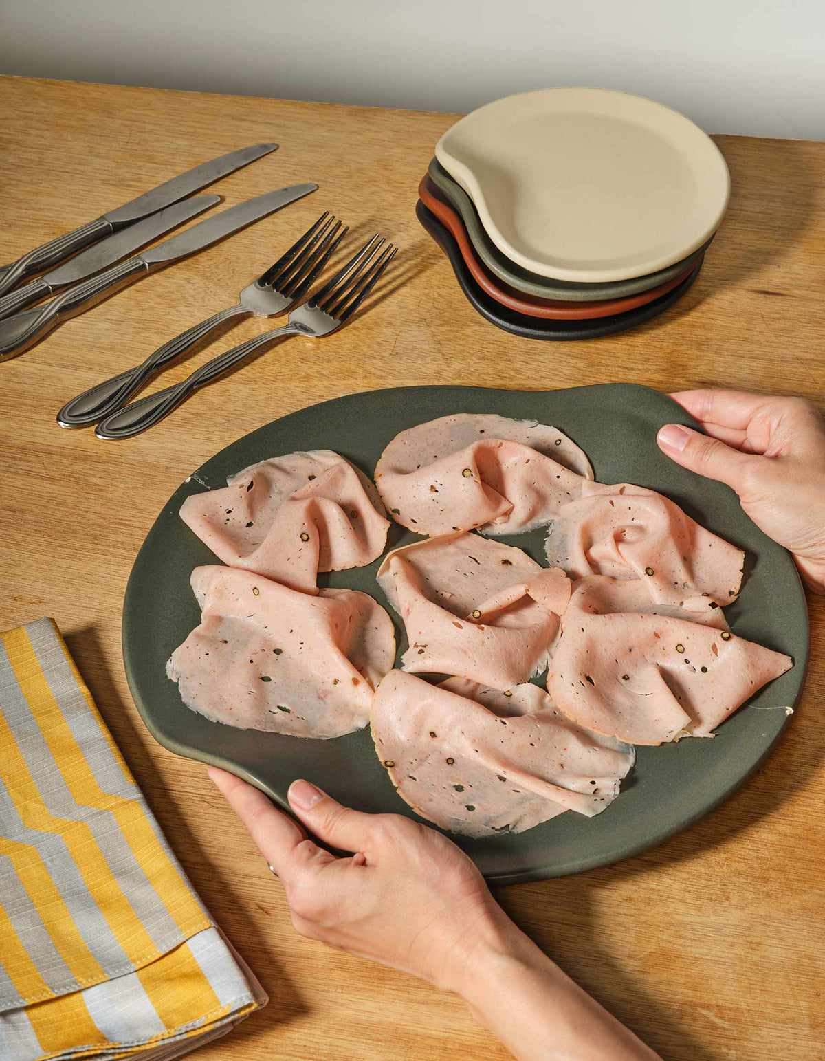 Two hands holding a Liche dark grey Tortuga Forma Serve Platter filled with folded pink sliced meat on a wooden dining table, beside cutlery, stacked plates and a yellow-and-white patterned napkin.