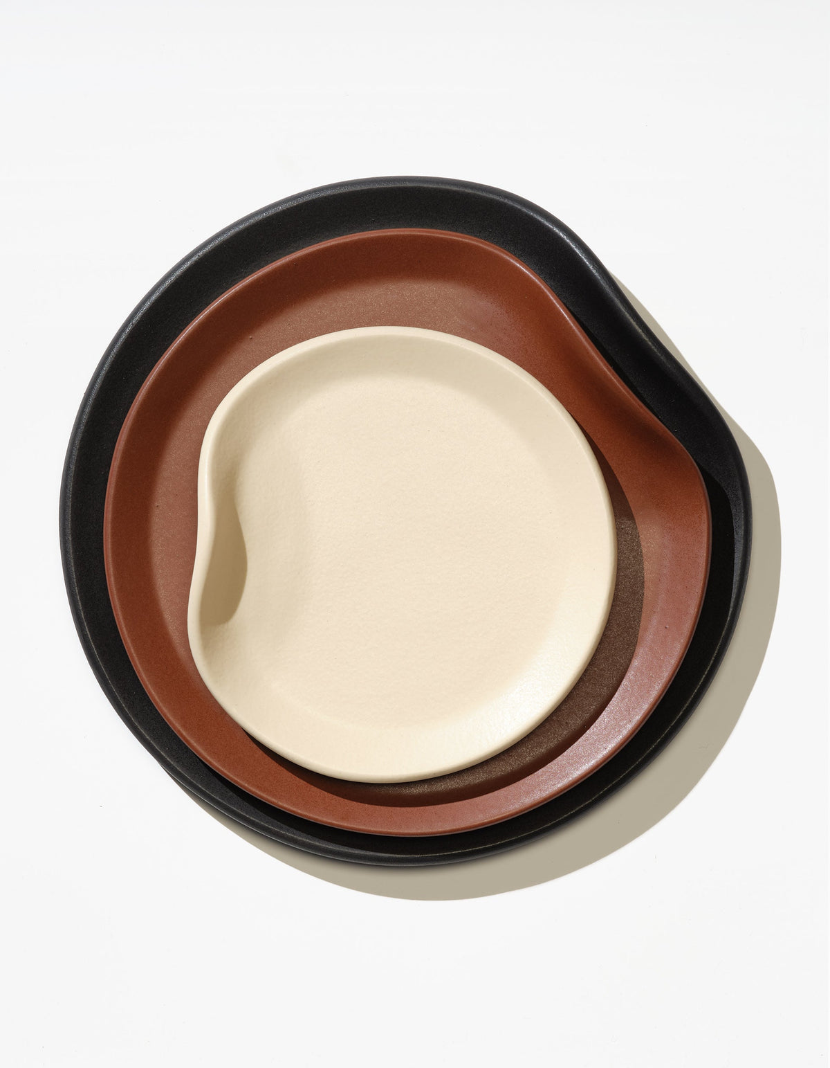 Tortuga Forma Dinner Plate Single in Soot beneath nested Tierra and Bone dishes, viewed overhead to show their contrasting colours, curved rims and irregular rounded shapes.