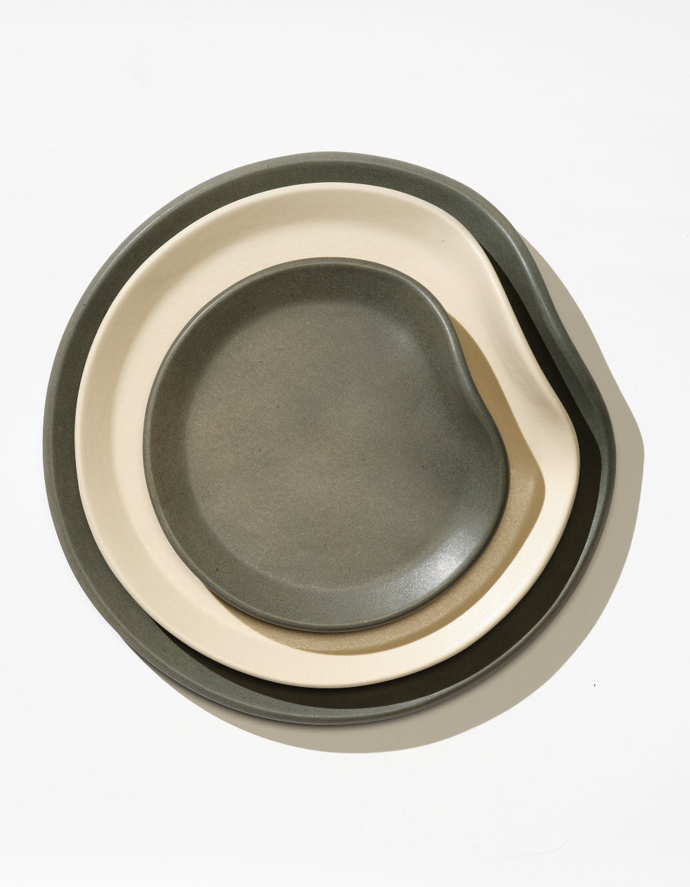 Tortuga Forma Bread Plate Single in Liche nested inside larger Bone and Liche plates, viewed overhead to show their contrasting finishes and matching irregular rounded outlines.