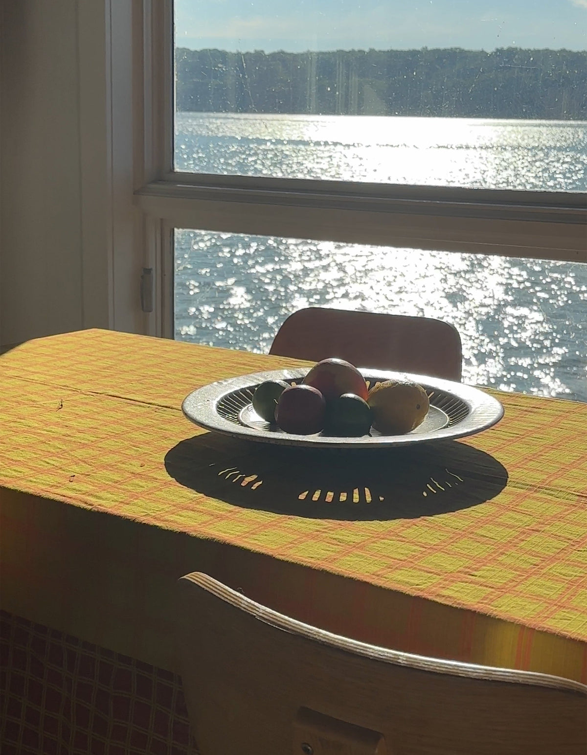 Mustard-yellow and pink Tortuga Forma Tablecloth Reversible Grid on a dining table beside a sunlit window overlooking water, with a shallow fruit bowl casting a strong shadow across the patterned fabric.