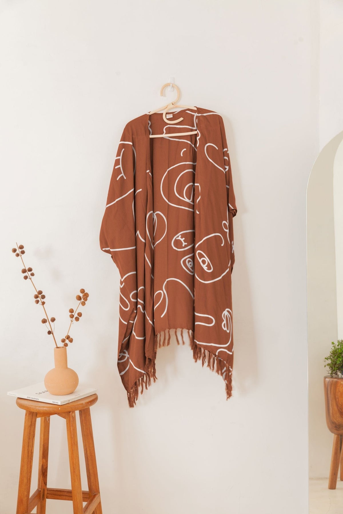 Village Thrive Faces Kimono, rust terracotta rayon with white line print, hanging on a wooden hanger against a wall.