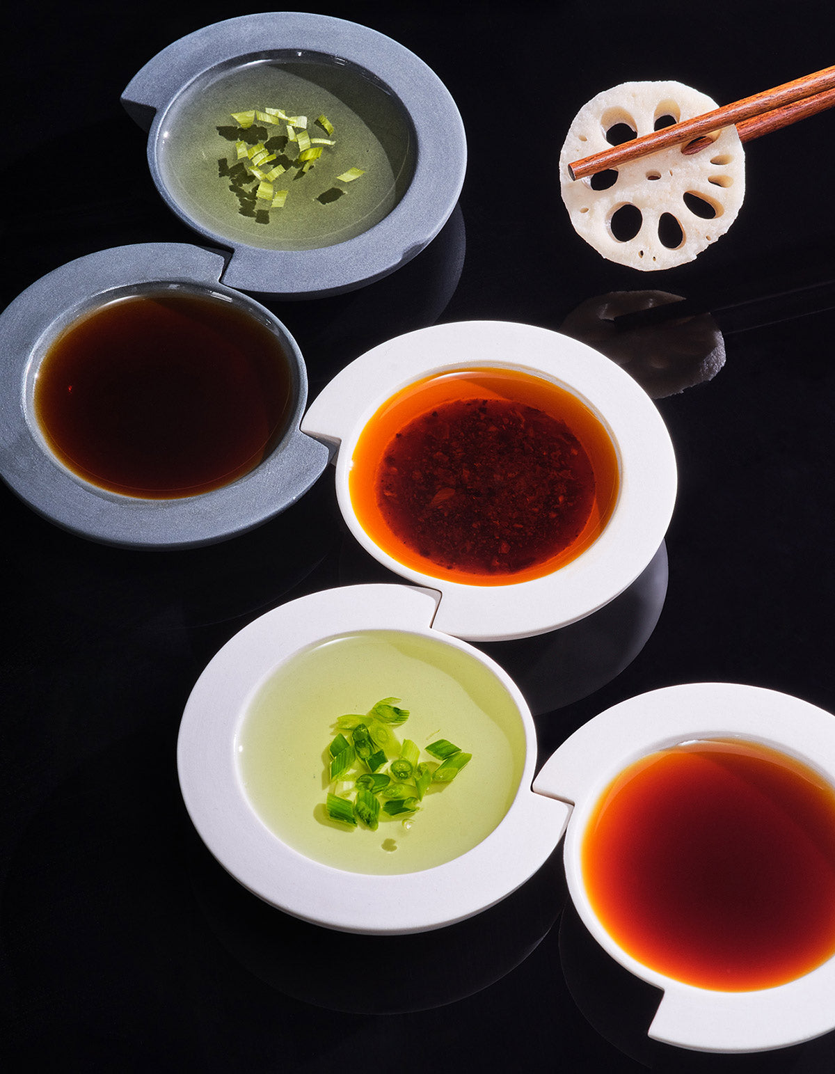 White and graphite Tortuga Forma Kaolin Sauce Dishes filled with dark brown, orange-red and pale green sauces on a glossy black surface, some topped with chopped green garnish, with chopsticks holding a lotus-root slice at the upper right.