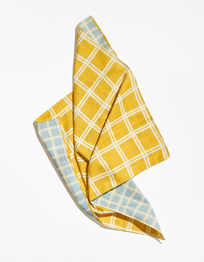 Tortuga Forma Dinner Napkins Double Sided Grid - High Season loosely folded on white, with a mustard-yellow and white grid face curling back to reveal a pale blue grid reverse and stitched border.