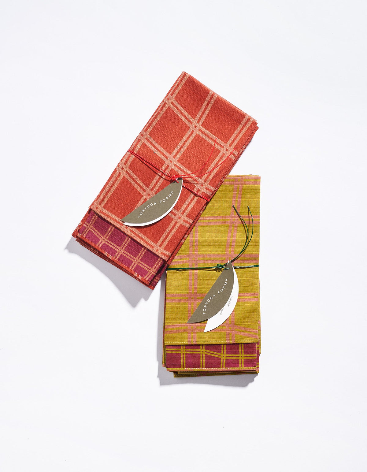 Two folded Tortuga Forma Dinner Napkins Double Sided Grid - High Season on white, one coral-red with a pale grid and the other mustard-yellow with a pink grid, each topped with a curved silver-coloured pendant on a dark cord.