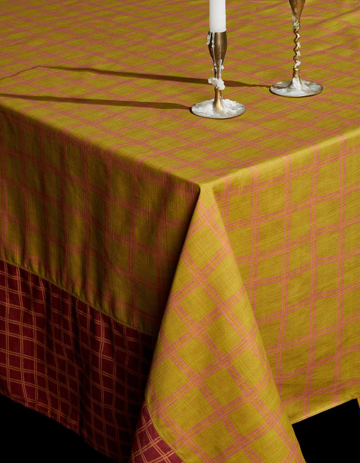 Tortuga Forma Tablecloth Reversible Grid draped over a table corner, showing a mustard-yellow face with a pink grid and a burgundy border with yellow grid lines, with two silver-coloured candlesticks on top.
