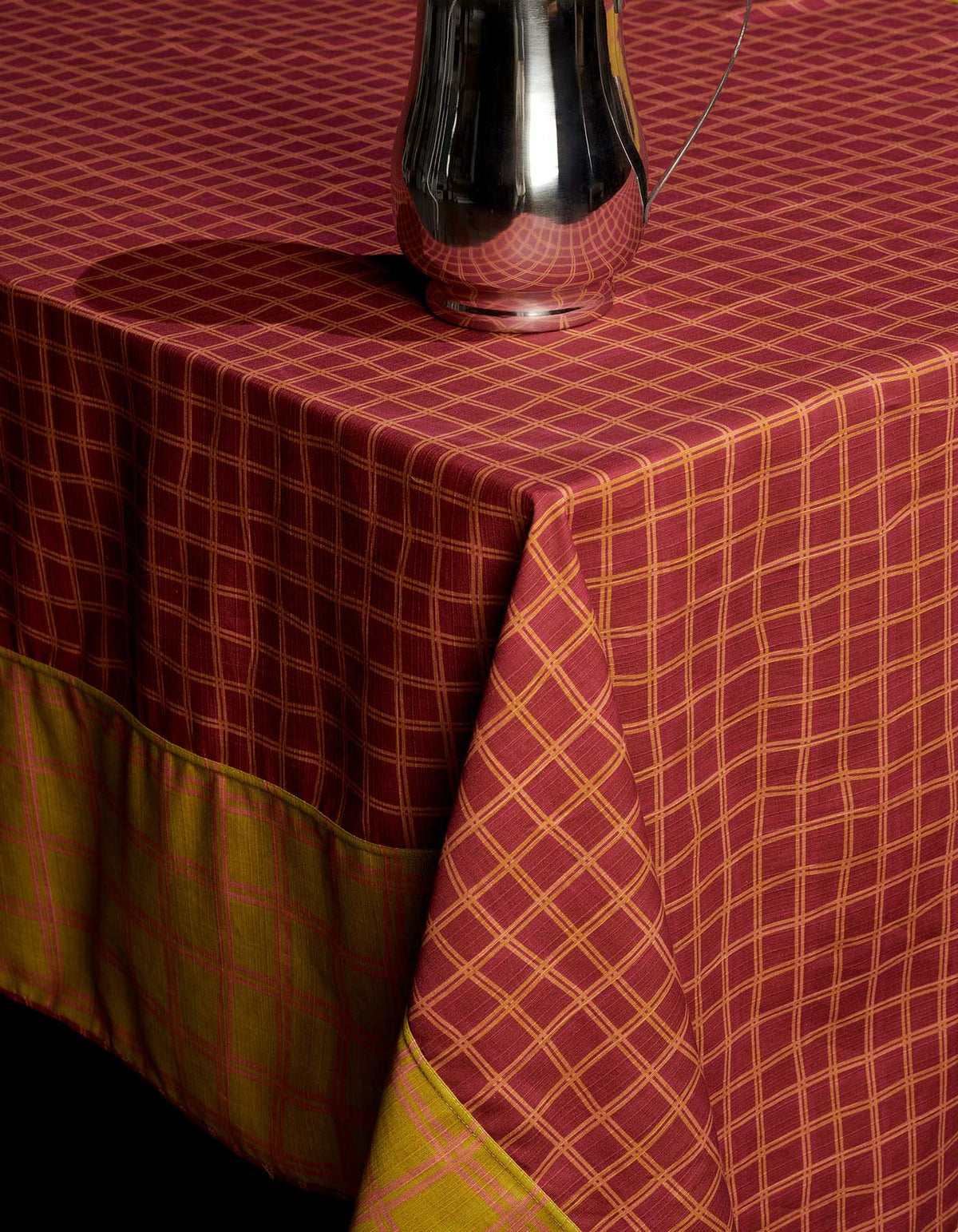 Burgundy side of Tortuga Forma Tablecloth Reversible Grid covering a table, showing a fine mustard-yellow grid and contrasting mustard border, with a reflective metal vessel above the folded corner.