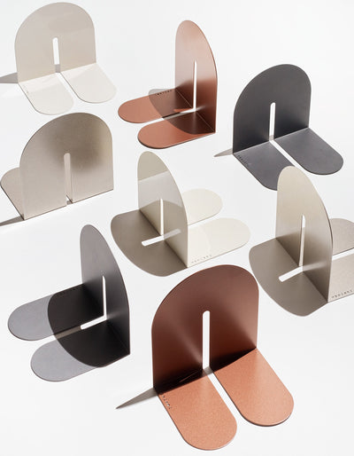 Tortuga Forma Dumbo Bookend Pairs - The Architect Collection arranged across a white surface in copper, cream, taupe and charcoal finishes, showing thin folded forms with arched upright panels, narrow central slots and rounded horizontal feet.