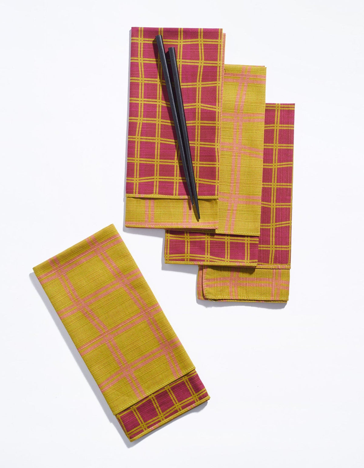 Tortuga Forma Dinner Napkins Double Sided Grid - High Season folded and overlapped on white, showing burgundy faces with mustard-yellow grids and mustard reverses with pink grids, with black chopsticks resting across the top napkin.