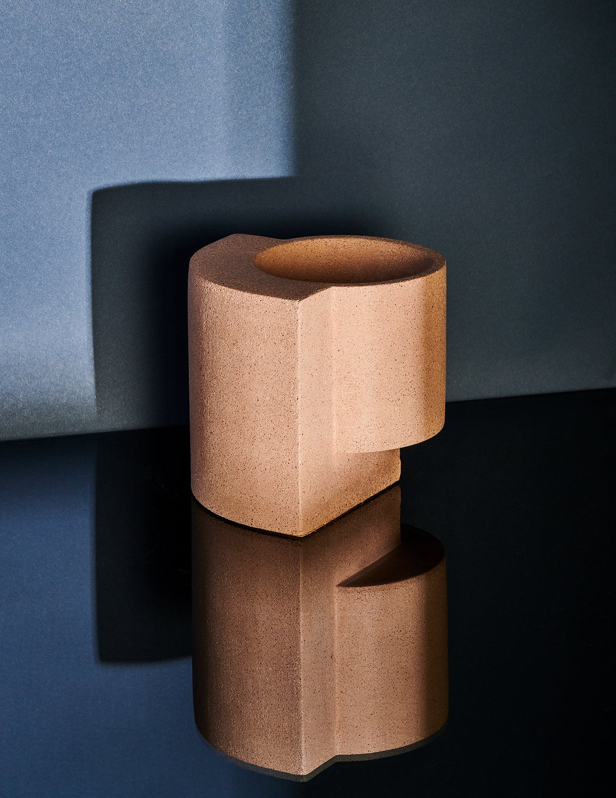 Coral Tortuga Forma Platform Planter Small - Terrazzo on a glossy black surface, casting a clear reflection against a blue-grey backdrop, with light highlighting its circular opening and geometric slab support.