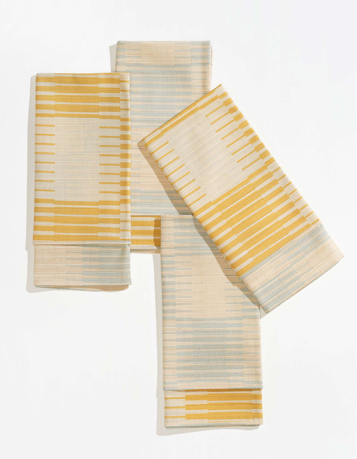Tortuga Forma Dinner Napkins Double Sided Drift - Quiet Season folded into overlapping rectangles on white, showing cream fabric patterned with mustard-yellow and pale grey-green lines of varying lengths, with neatly stitched borders.