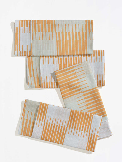 Four folded Tortuga Forma Dinner Napkins Double Sided Drift - Quiet Season arranged on white, showing orange and pale grey linear patterns forming staggered bands across the fabric.