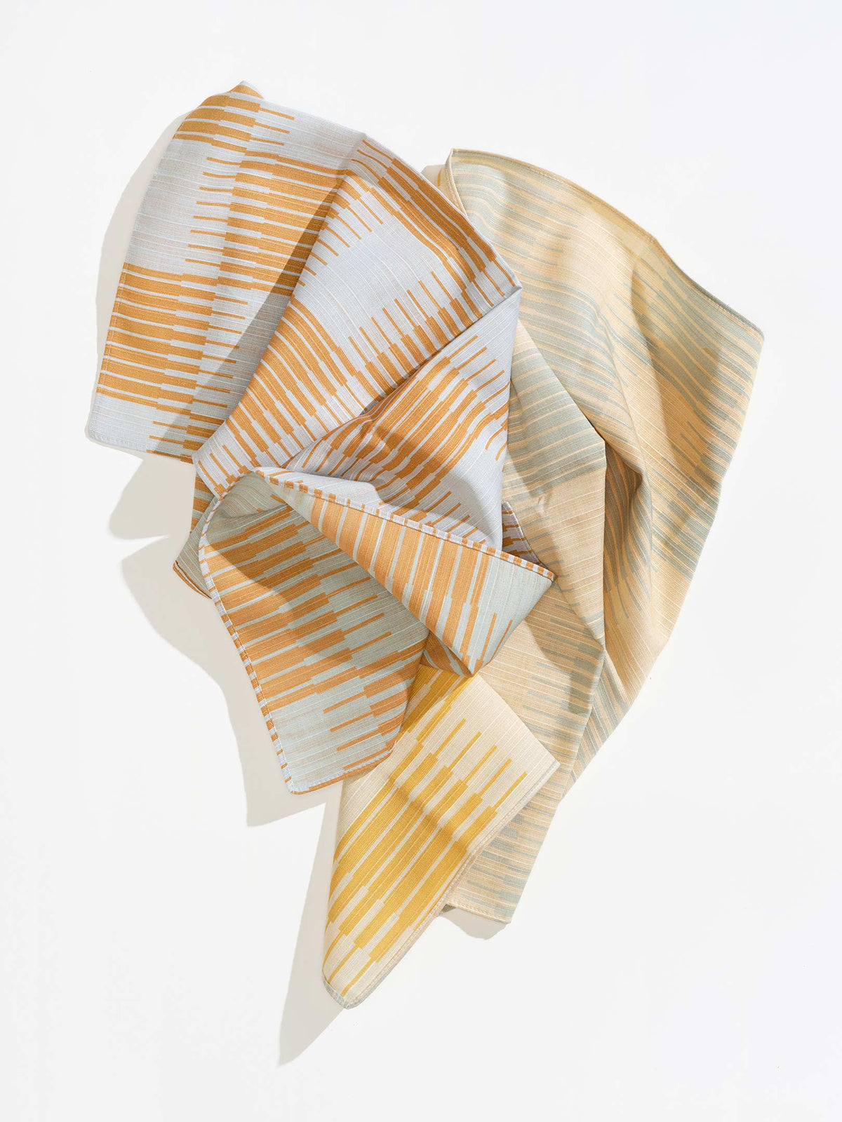 Loosely draped Tortuga Forma Dinner Napkins Double Sided Drift - Quiet Season on white, showing overlapping folds of cream fabric with orange, mustard-yellow and pale grey-green lines and stitched edges.