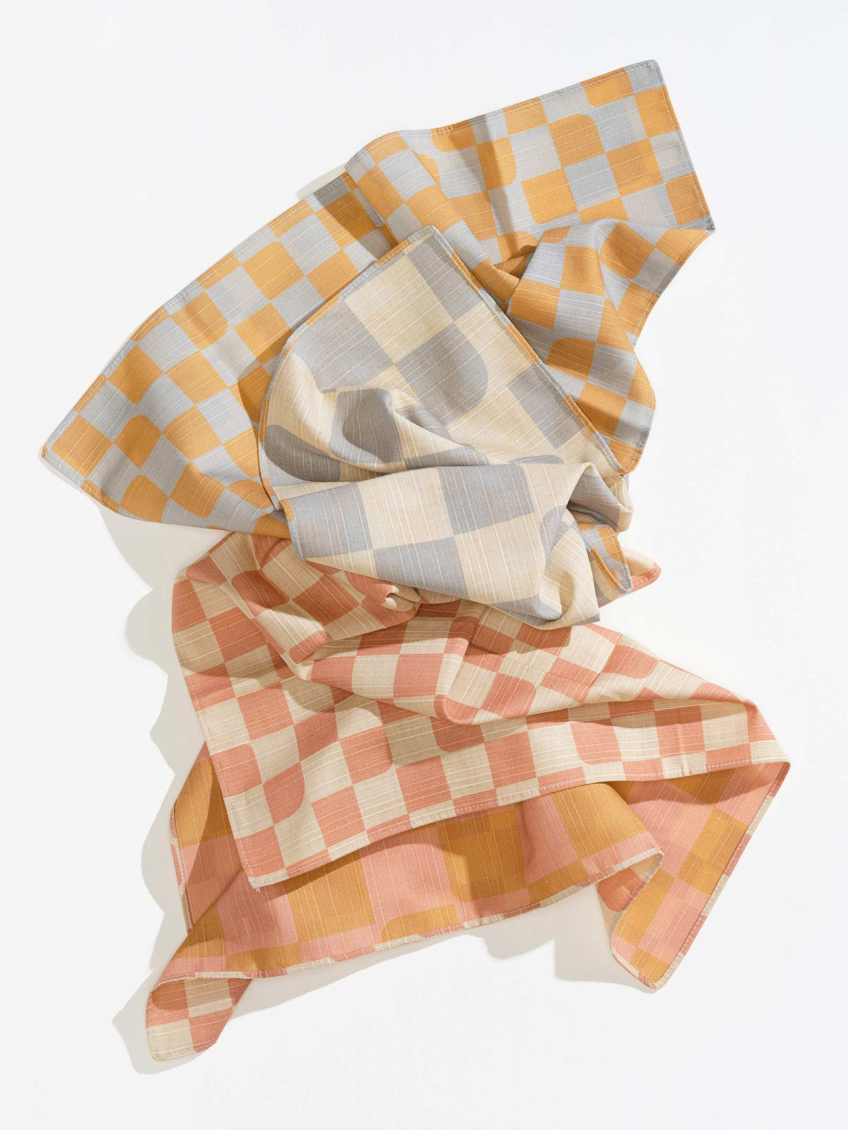 Tortuga Forma Dinner Napkins Double Sided Checker - Quiet Season loosely draped on white, showing mustard-and-grey, cream-and-grey, coral-and-cream and pink-and-mustard checker patterns across overlapping folds.