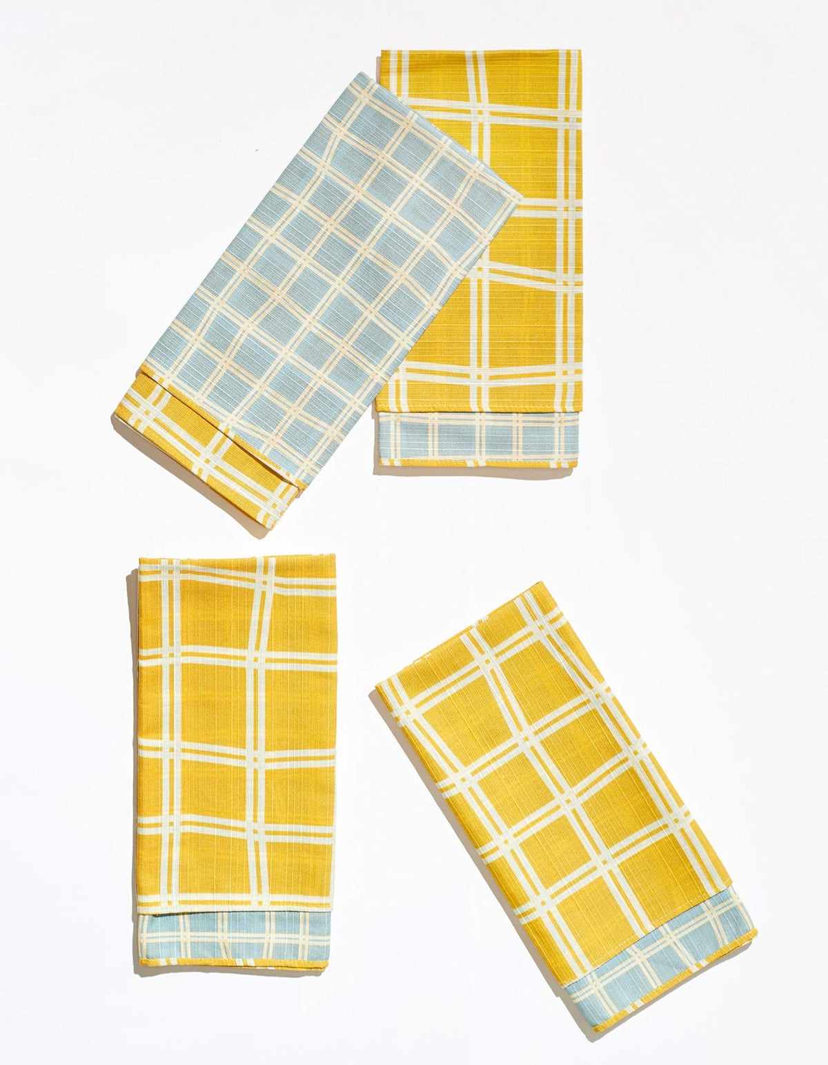 Four folded Tortuga Forma Dinner Napkins Double Sided Grid - High Season arranged on white, showing mustard-yellow faces with white grids and pale blue reverses with yellow-and-white grids.