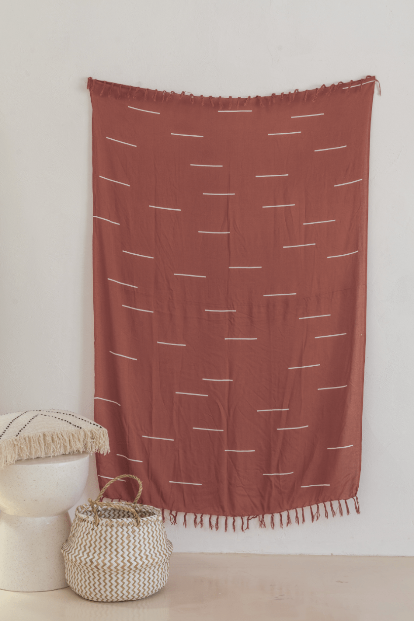 Village Thrive Lines Tapestry, rust terracotta rayon with white line print, hanging above a woven basket.