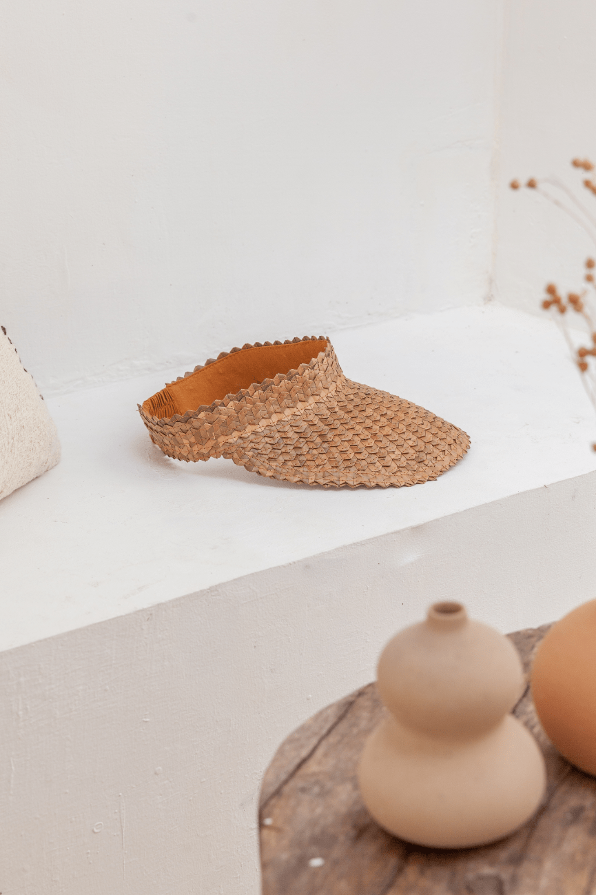 Village Thrive Rattan Sun Visor, amber palm leaf, displayed on a shelf beside ceramic pottery.