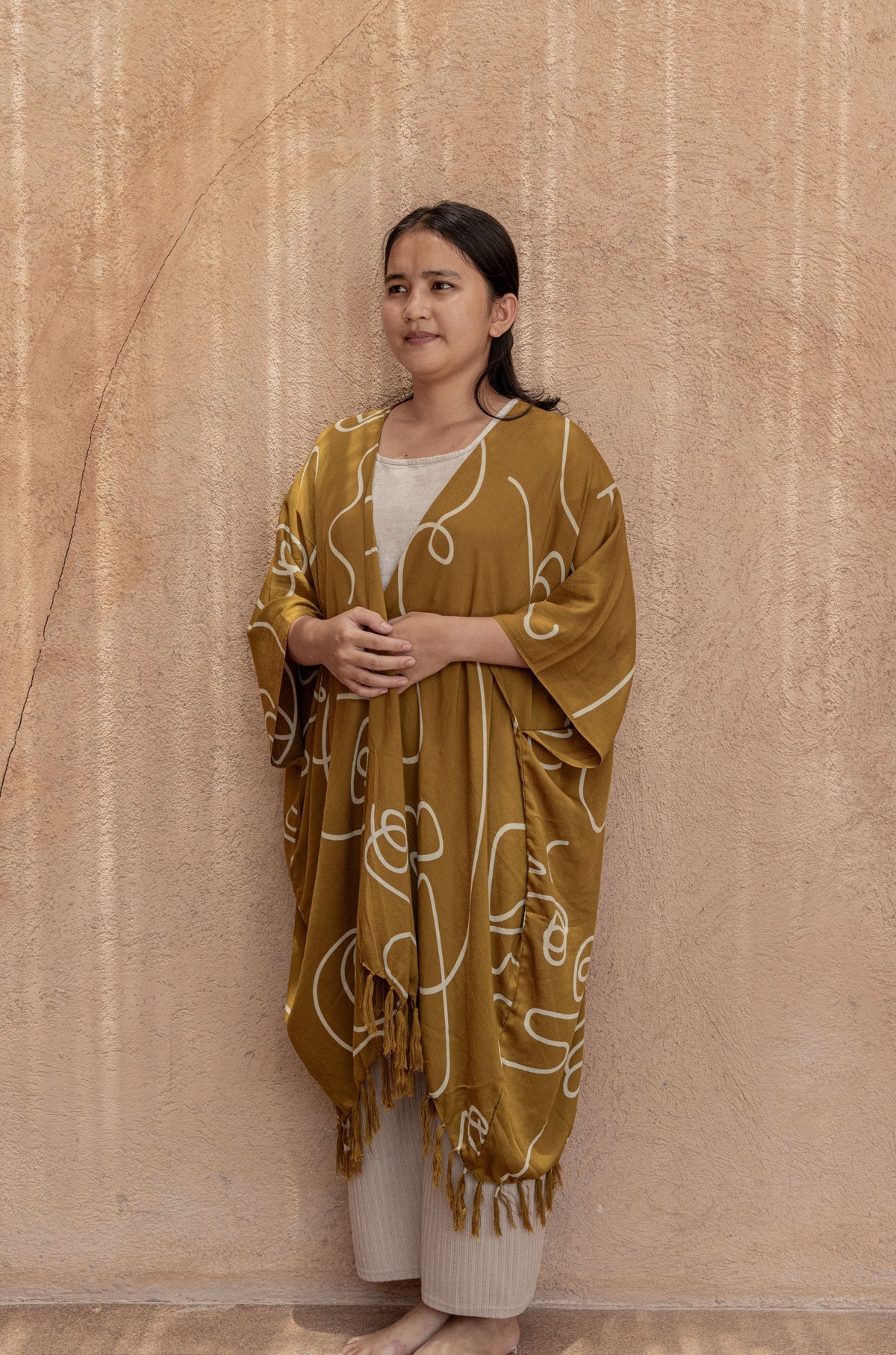 Village Thrive Faces Kimono, olive gold rayon, worn by a model standing against a wall.